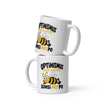 Weiße Keramiktasse mit lustigem Bienenmotiv und dem Spruch „Optimismus heißt umgekehrt Sumsi mit Po“ in schwarzer und gelber Schrift.