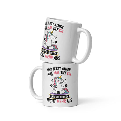 Weiße Keramiktasse mit einem meditierenden Einhorn und dem lustigen Spruch „Und jetzt atmen alle mal tief ein und die Idioten nicht mehr aus“ in schwarzer und pinker Schrift.